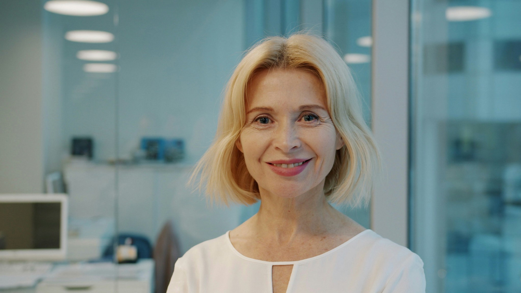 A smiling blonde woman in a white shirt indoors.