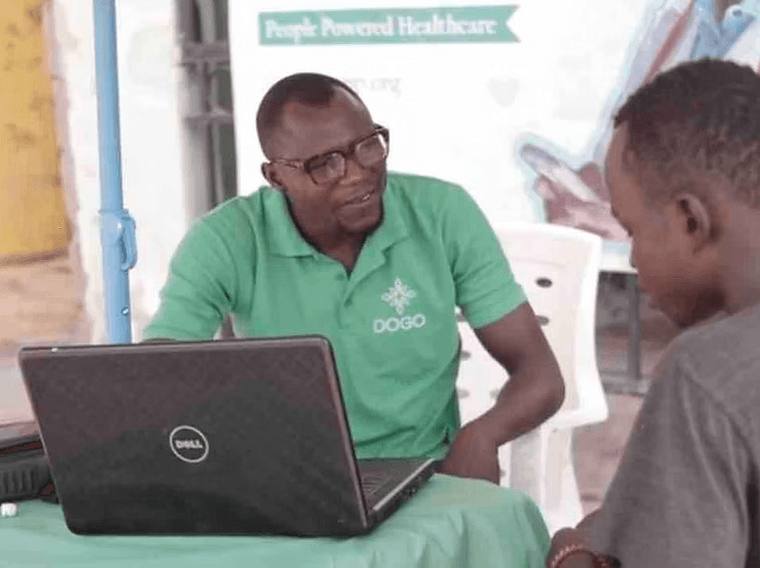 A DOGO team member in a branded polo shirt registering a visitor at a community stand beneath a People Powered Healthcare banner.